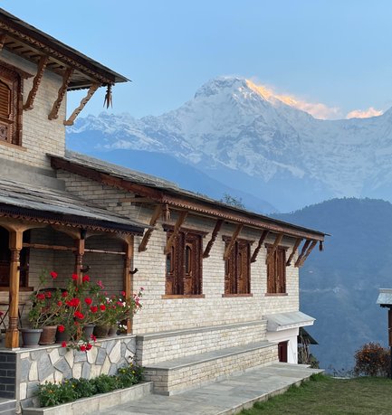Ein traditionelles Haus mit kunstvollen Holzfenstern steht vor majestätischen, schneebedeckten Bergen unter einem klaren Himmel.