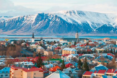 Blick auf Reykjavik mit bunten Häusern, umgeben von schneebedeckten Bergen und einem ruhigen Wasser im Hintergrund.