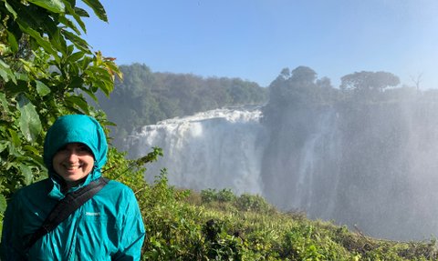 Eine Person in einer blauen Regenjacke lächelt vor den beeindruckenden Victoriafällen, umgeben von üppigem Grün.