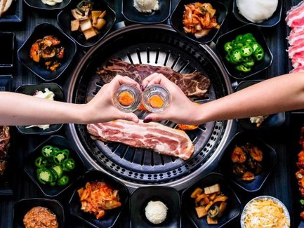 Zwei Hände halten Gläser mit einem orangefarbenen Getränk, umgeben von verschiedenen koreanischen BBQ-Gerichten auf einem Tisch.