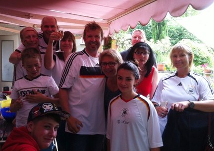 Eine fröhliche Gruppe von Menschen in Fußballtrikots feiert zusammen unter einem Vordach, umgeben von Pflanzen und Getränken.