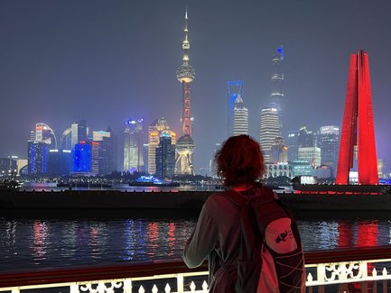 Eine Person mit lockigem Haar steht an einem Geländer und blickt auf die beleuchtete Skyline von Shanghai bei Nacht.