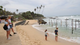 Am Strand stehen zwei Kinder und eine Frau, während die Wellen sanft an den Sand schlagen und Palmen im Hintergrund wehen.
