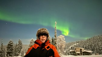 Eine Person steht in einem verschneiten Landschaftsbild, während die Nordlichter in lebhaften Grüntönen am Himmel leuchten.