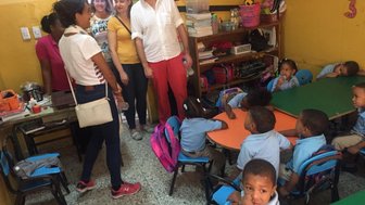 Eine Gruppe von Erwachsenen interagiert mit Kindern in einem Klassenzimmer, das mit bunten Tischen und Stühlen ausgestattet ist.