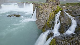 Der majestätische Wasserfall fließt über grüne Felsen, umgeben von einer malerischen Landschaft mit sanften Hügeln im Hintergrund.