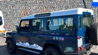 Ein blauer Geländewagen steht auf einem Parkplatz, umgeben von grünem Gras und einem steinernen Hintergrund.