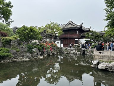 Ein traditionelles chinesisches Gartenhaus mit geschwungenem Dach, umgeben von üppigem Grün und bunten Blumen am Teich.
