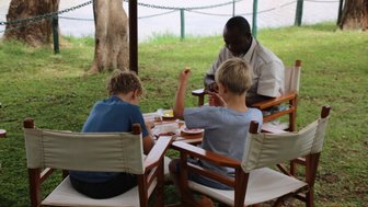 Zwei Kinder sitzen an einem Tisch im Freien und genießen eine Mahlzeit, während ein Erwachsener ihnen zuschaut.