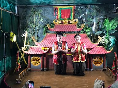 Zwei Puppenspieler stehen in knalligen Kostümen vor einer traditionellen Kulisse, umgeben von Wasser und Pflanzen.