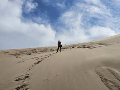 Ein Mensch erklimmt eine hohe Sanddüne in der Mongolei, umgeben von einem weiten, wolkigen Himmel.