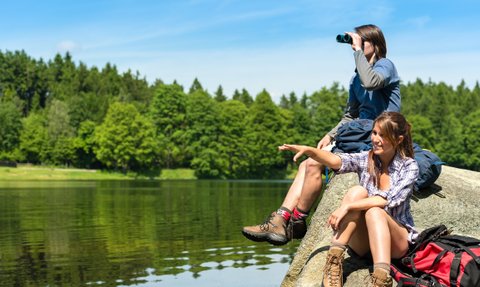 Zwei Jugendliche sitzen auf einem Felsen am Wasser, während einer mit einem Fernglas in die Ferne schaut.