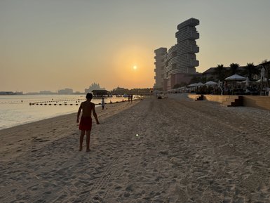 Ein Junge in roten Shorts geht am Strand entlang, während die Sonne hinter modernen Gebäuden untergeht.