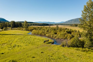 Eine Gruppe von Reitern durchquert einen ruhigen Fluss, umgeben von üppigem Grün und majestätischen Bergen im Hintergrund.