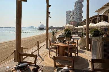 Ein elegantes Strandrestaurant mit Holztischen und Stühlen, umgeben von Palmen und Sonnenschirmen, bietet einen Blick auf das Wasser.