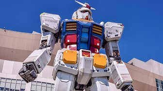Ein riesiger Gundam-Roboter steht majestätisch vor einem modernen Gebäude in Tokyo, umgeben von klarem blauen Himmel.