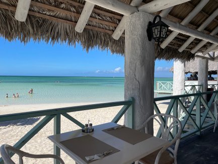 Ein Blick auf einen Strand mit klarem Wasser, wo Kinder im seichten Wasser spielen, während ein Restaurant im Hintergrund sichtbar ist.