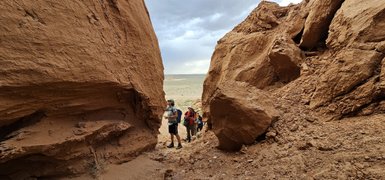 Drei Wanderer erkunden eine beeindruckende Felslandschaft mit großen, rotbraunen Felsen und einer weiten, offenen Ebene im Hintergrund.
