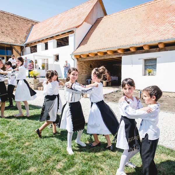 Eine Gruppe von Kindern tanzt fröhlich im Freien, gekleidet in traditionelle Trachten, vor einem ländlichen Hintergrund.