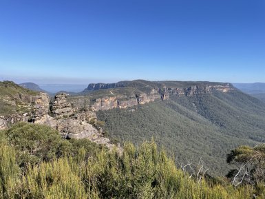 Ein atemberaubender Blick auf die felsigen Klippen der Blue Mountains, umgeben von üppiger grüner Vegetation und strahlend blauem Himmel.