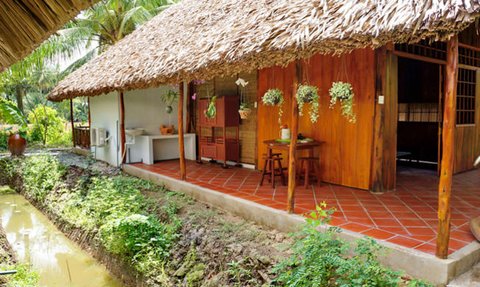 Ein traditionelles, strohgedecktes Haus mit Holzverkleidung, umgeben von üppigem Grün und einem kleinen Wasserlauf.