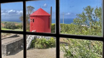 Durch ein Fenster sieht man einen roten Turm, umgeben von üppigem Grün und dem blauen Ozean im Hintergrund.