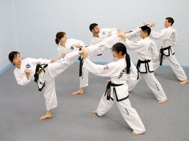 Sechs Taekwondo-Praktizierende in weißen Anzügen üben gemeinsam Techniken in einem hellblauen Raum.