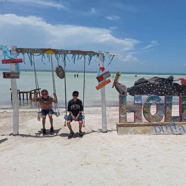 Zwei Kinder schwingen fröhlich auf einer Schaukel am Strand von Holbox, umgeben von klarem Wasser und einem blauen Himmel.