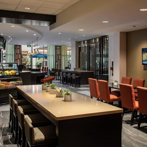 Ein modernes Café mit einer eleganten Theke, umgeben von gemütlichen Stühlen und Pflanzen auf den Tischen.