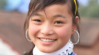 Ein fröhliches vietnamesisches Mädchen mit langen Haaren und großen Ohrringen lächelt in die Kamera, umgeben von einer ländlichen Kulisse.