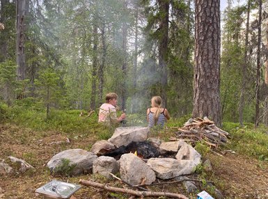 Zwei Personen sitzen am Lagerfeuer in einem Wald, umgeben von hohen Bäumen und einer ruhigen, natürlichen Umgebung.