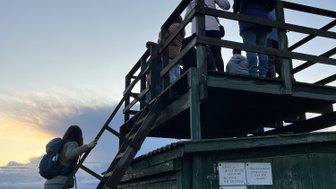 Eine Gruppe von Menschen steht auf einer Aussichtsplattform und beobachtet den Sonnenuntergang über einer Landschaft.