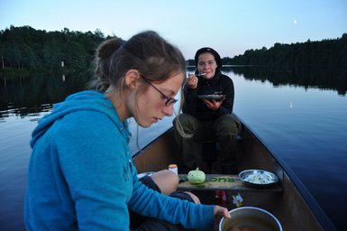 Zwei Teenager sitzen in einem Kanu auf einem ruhigen See, während sie eine Mahlzeit genießen und die Natur um sich herum bewundern.