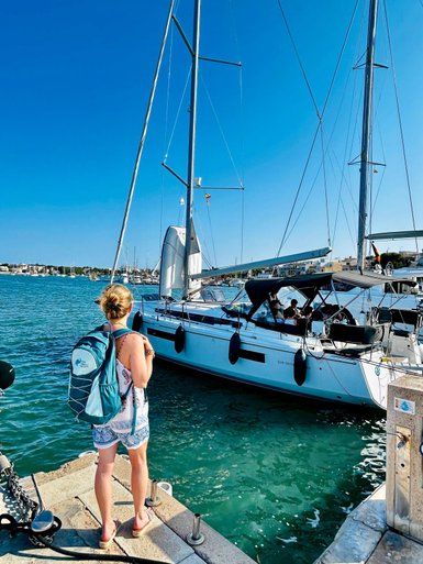 Eine Frau mit einem Rucksack steht am Hafen und beobachtet ein Boot, das im klaren Wasser anlegt.