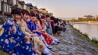 Eine Gruppe junger Frauen in farbenfrohen Kimonos sitzt am Ufer eines Flusses in Kyoto und genießt die Abendstimmung.