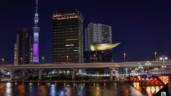 Die Tokyo Skytree leuchtet in bunten Farben, während moderne Gebäude und eine Brücke den nächtlichen Fluss säumen.
