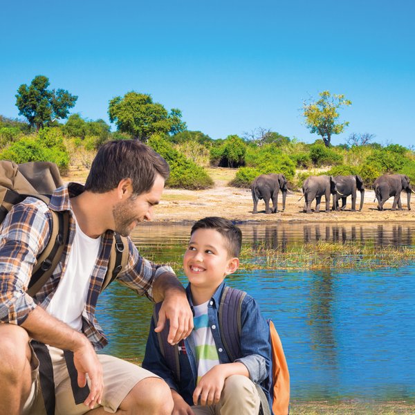 Ein Vater und sein Sohn lächeln sich am Ufer eines Gewässers an, während Elefanten im Hintergrund grasen.