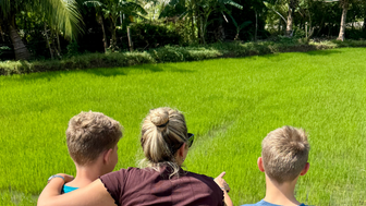 Eine Frau zeigt auf ein grünes Reisfeld, während sie mit zwei Jungen in der Natur steht, umgeben von Palmen.
