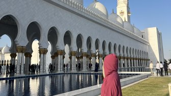 Eine Person in einem roten Gewand steht am Rand eines Wasserspiegels, während die beeindruckende Sheikh Zayed Moschee im Hintergrund sichtbar ist.