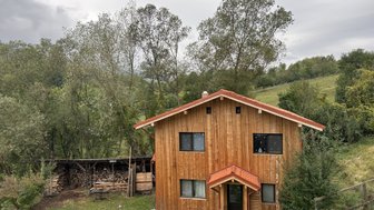 Ein rustikales Holzhaus mit rotem Dach steht umgeben von Bäumen und einer grünen Wiese, die unter einem bewölkten Himmel liegt.