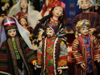 Drei kunstvoll gestaltete Puppen in traditioneller Kleidung stehen vor einem bunten Hintergrund, umgeben von weiteren Puppen.