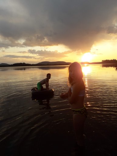 Zwei Kinder genießen den Sonnenuntergang am Wasser, während eines auf einem Reifen sitzt und das andere im Wasser steht.