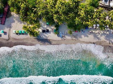 Eine luftige Perspektive zeigt den langen, ruhigen Strand mit palmengesäumter Küste und den sanft brechenden Wellen.