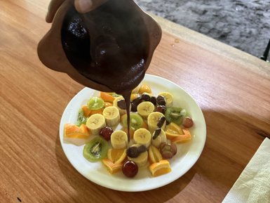 Eine Hand gießt dicke Schokoladensauce über einen bunten Obstteller mit Bananen, Trauben und Kiwi.