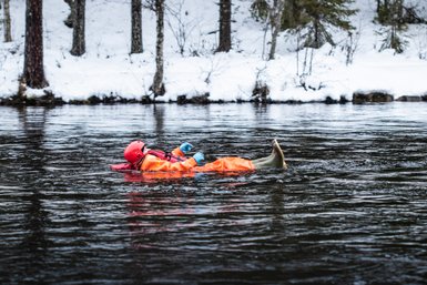 Eine Person in einem orangefarbenen Anzug schwimmt entspannt auf einem ruhigen Fluss, umgeben von schneebedeckten Bäumen.