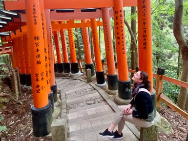 Eine Person sitzt auf einer Steinbank und blickt lächelnd zu den leuchtend orangefarbenen Torii-Toren auf einem schmalen Weg.