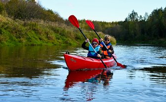 Zwei Kinder paddeln in einem roten Kajak auf einem ruhigen Fluss, umgeben von grünen Ufern und Bäumen im Hintergrund.