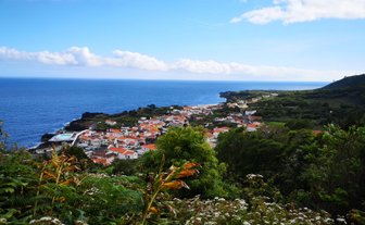 Ein malerischer Blick auf ein Küstendorf mit roten Dächern, umgeben von üppigem Grün und dem blauen Ozean im Hintergrund.