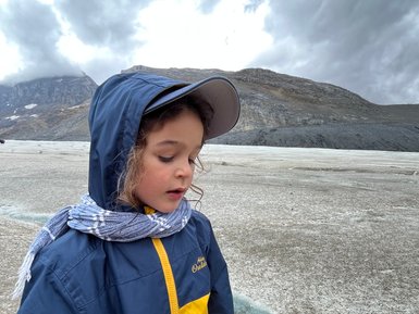 Ein Kind in einem blauen und gelben Regenjacke steht auf einem Gletscher, umgeben von grauen Wolken und Bergen.