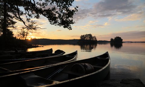 Am Ufer eines ruhigen Sees liegen mehrere Kanus, während die Sonne hinter den Bäumen untergeht und den Himmel erleuchtet.